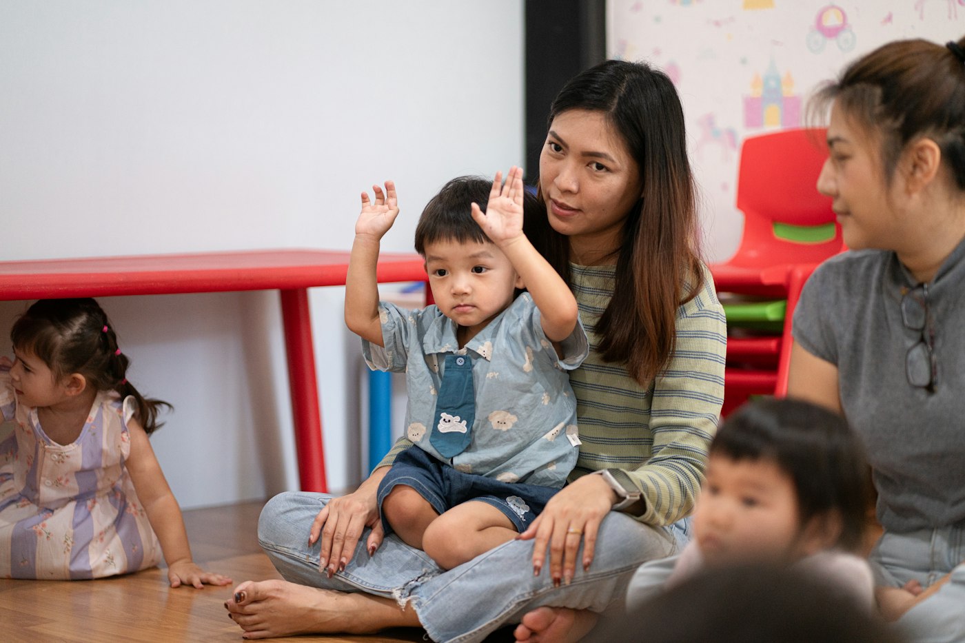 A practitioner sitting on the floor at group time with a toddler on her lap, the child raising both hands