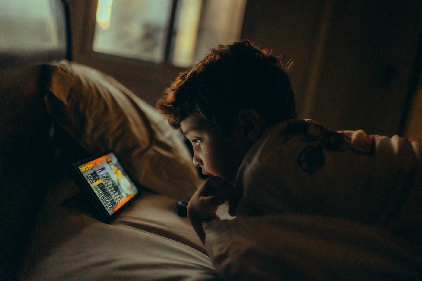 A young child lying in bed in a dark room, looking at a tablet