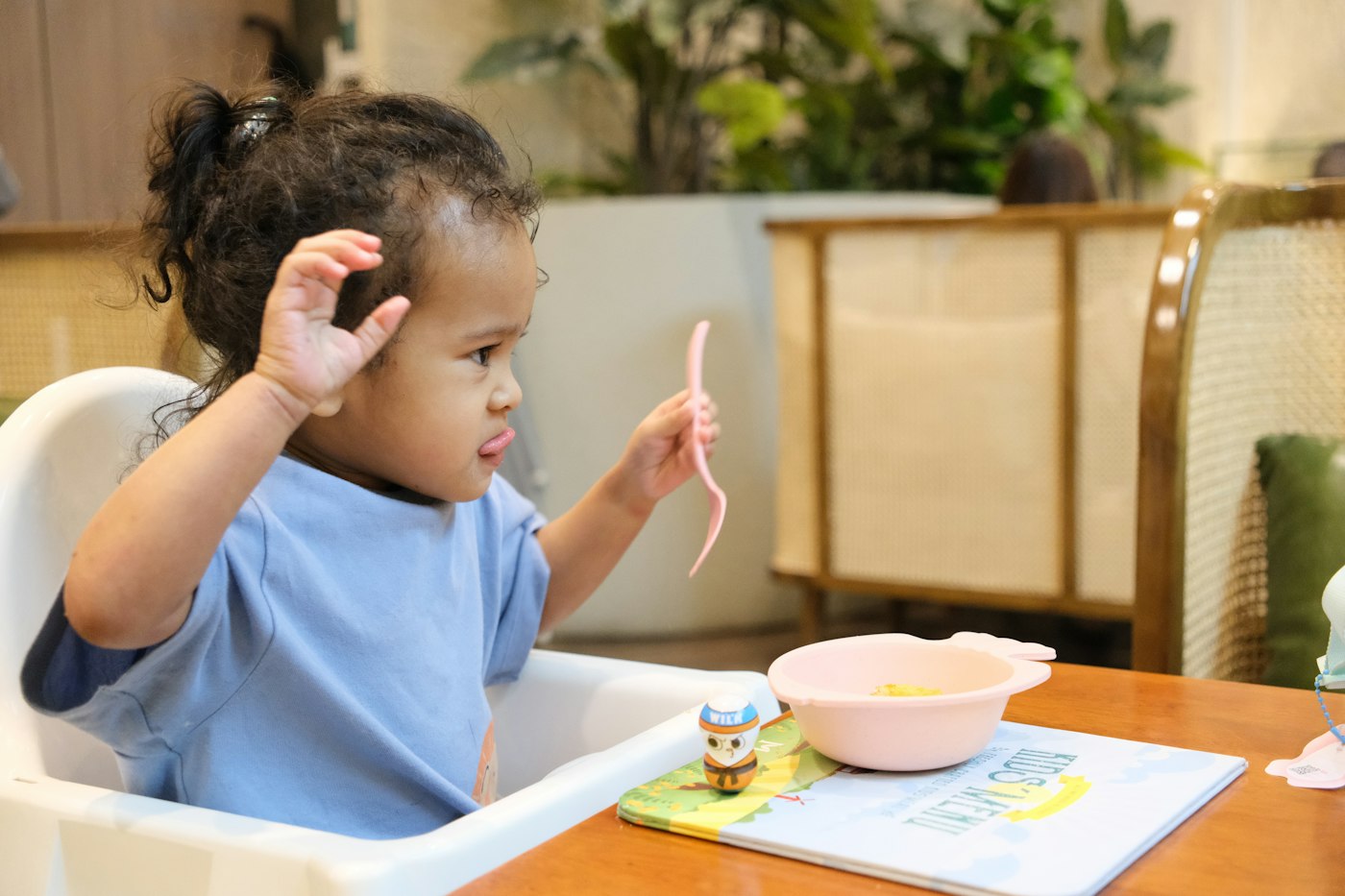 A toddler in a high chair feeding themselves with a spoon and bowl